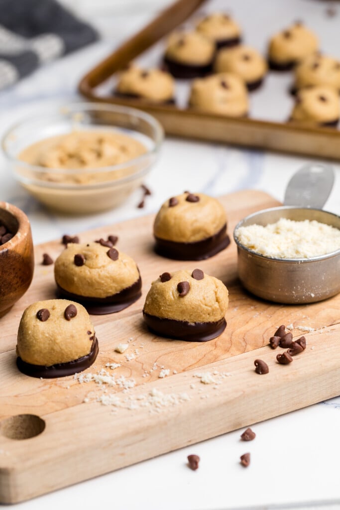 Three balls of cookie dough dipped in chocolate and topped with chocolate chips rest on a wooden board, with measuring cups of flour and chocolate chips and more dough balls on a tray in the background.