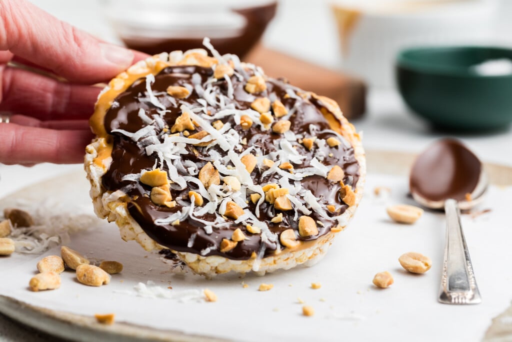 A hand holds a Chocolate Peanut Butter Rice Cake Treat topped with chocolate, shredded coconut, and chopped peanuts. The rice cake rests on parchment paper with scattered peanuts and a spoon nearby.
