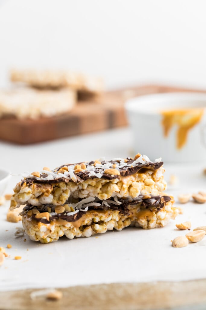Two stacked Chocolate Peanut Butter Rice Cake Treats with a chocolate coating and sprinkled coconut, filled with peanut butter, rest on white parchment paper, surrounded by peanuts, with a blurred cup and cutting board in the background.