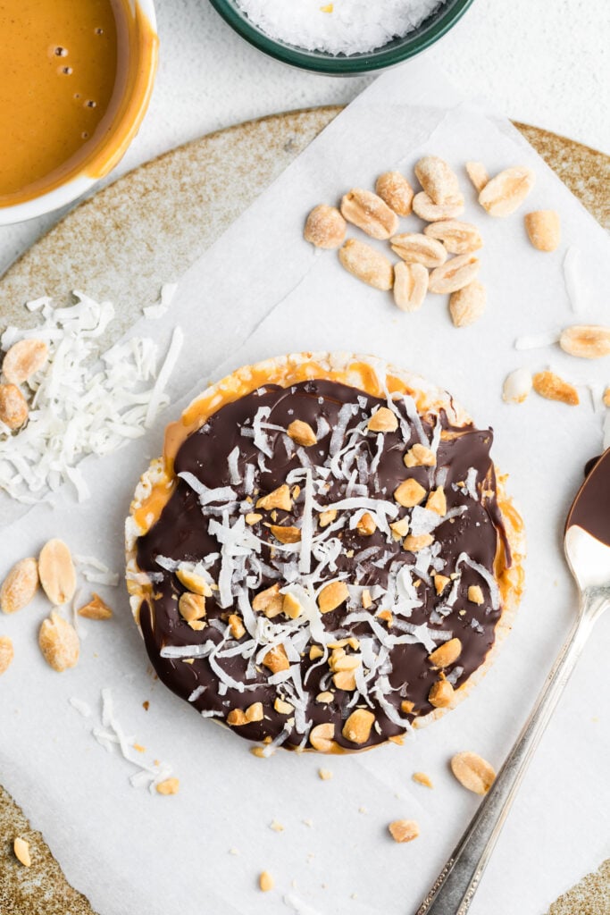 A Chocolate Peanut Butter Rice Cake Treat topped with melted chocolate, shredded coconut, and chopped peanuts sits on parchment paper, surrounded by a spoon, peanuts, shredded coconut, and a bowl of peanut butter.