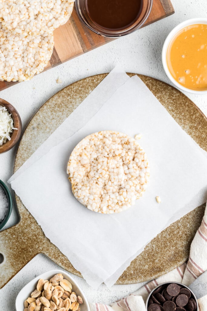 A plain rice cake sits on parchment paper atop a round board, surrounded by small bowls of chocolate, peanut butter, peanuts, shredded coconut, and chocolate chips—perfect for making Chocolate Peanut Butter Rice Cake Treats.