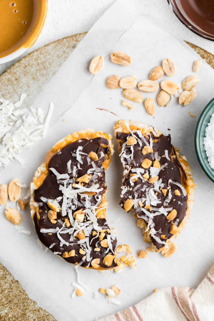 A Chocolate Peanut Butter Rice Cake Treat is broken in half on parchment paper, topped with chocolate, shredded coconut, and peanuts, and surrounded by loose coconut, peanuts, and a bowl of salt.