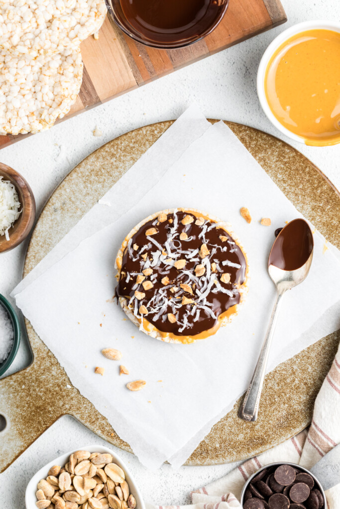 A Chocolate Peanut Butter Rice Cake Treat sits on parchment paper, topped with chocolate, shredded coconut, and chopped peanuts—surrounded by a spoon and bowls of peanut butter, coconut, chocolate, and peanuts.