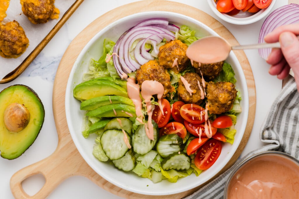 A salad bowl with lettuce, sliced avocado, cucumber, grape tomatoes, red onion, and meatballs, being drizzled with creamy dressing. There is a halved avocado and a bowl of cherry tomatoes nearby.