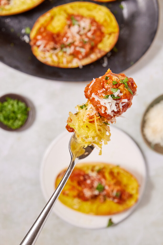 A fork holds spaghetti squash topped with tomato sauce and grated cheese, accompanied by savory turkey meatballs with spaghetti squash, with a stuffed squash half and bowls of herbs and cheese in the background.