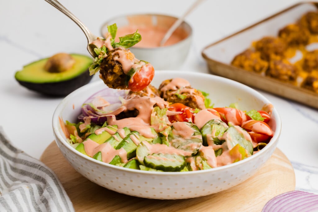 A bowl filled with fresh vegetables, chopped cucumbers, tomatoes, red onions and falafel pieces, all drizzled with creamy pink sauce. A fork picks up a morsel from the bowl. There are an avocado and sauce in the background.