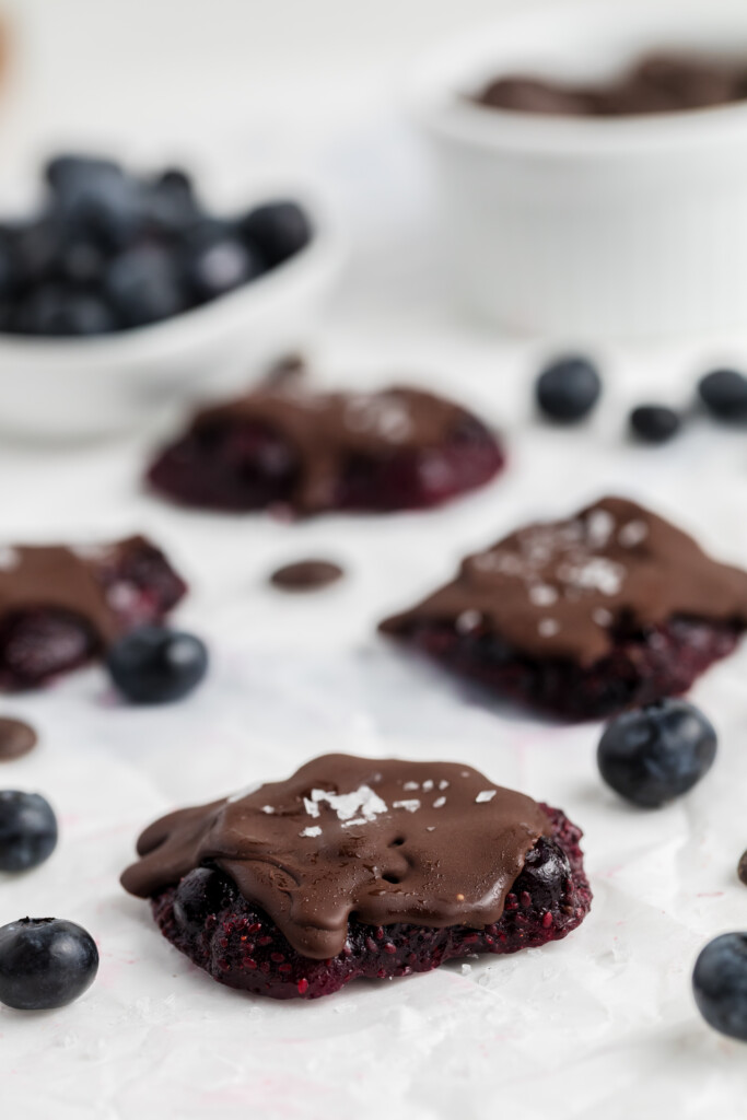 Close-up of chocolate-covered blueberry clusters on parchment paper, sprinkled with flaky salt. Fresh blueberries and a white bowl filled with blueberries are blurred in the background.