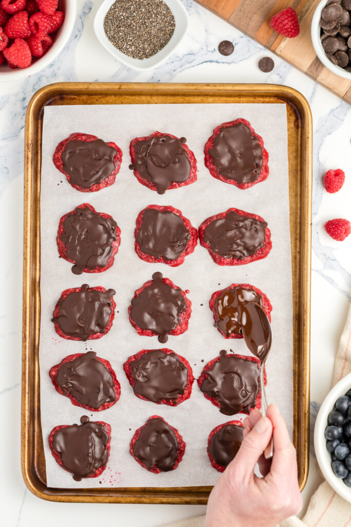 A hand spooning melted chocolate onto raspberry cookies arranged on a baking sheet lined with parchment paper. Bowls of raspberries, chia seeds, and chocolate chips are visible nearby.