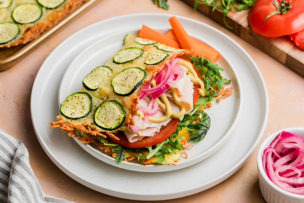 A veggie sandwich with zucchini, tomato slices, leafy vegetables, pickled onions and carrot sticks, placed on a zucchini cheese flatbread base, served on a white plate with fresh vegetables in the background.