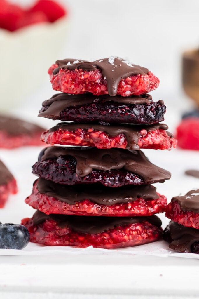A stack of red and dark chocolate-dipped cookies is surrounded by fresh raspberries and blueberries. Some cookies are topped with sea salt, and bowls of berries and chocolate are blurred in the background.