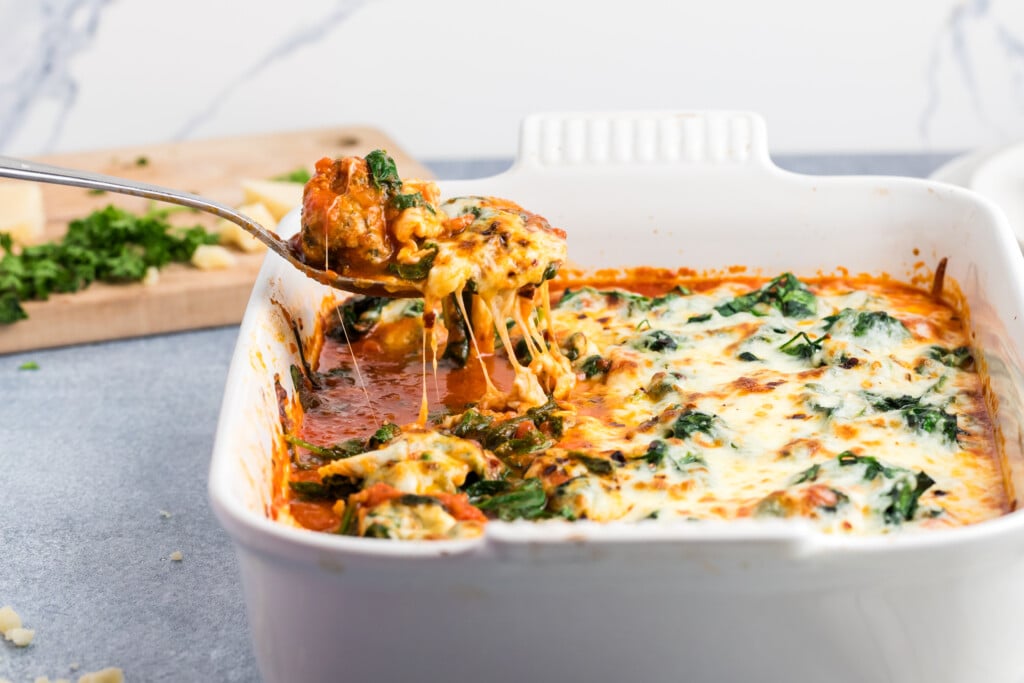A spoon lifts a serving of baked cheesy spinach and tomato casserole from a white baking dish, with melted cheese stretching. Chopped herbs and a cutting board are blurred in the background.