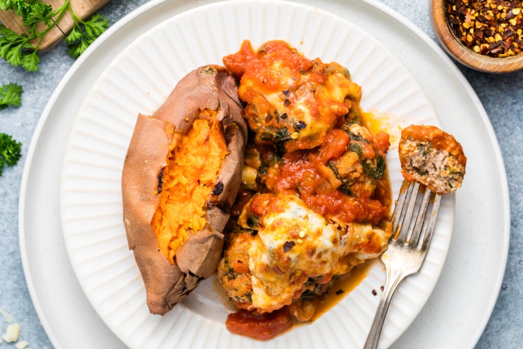 A plate with a baked sweet potato and cheesy meatballs topped with tomato sauce. A fork with a piece of meatball is on the plate, and a small bowl of red pepper flakes is nearby.