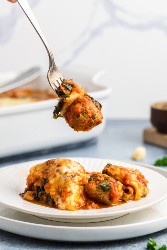 A fork holds a portion of cheesy, baked stuffed shells covered in tomato sauce above a white plate with more stuffed shells, with a baking dish in the background.