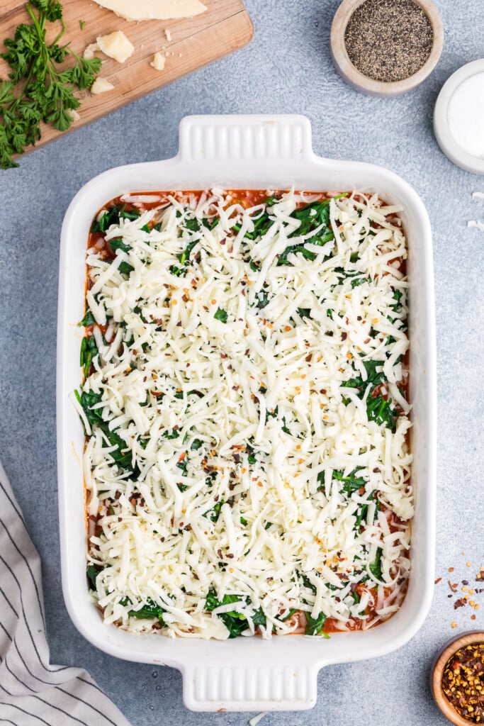 A rectangular white baking dish filled with an uncooked lasagna topped with shredded cheese and spinach. Nearby are bowls of pepper, salt, crushed red pepper, a striped towel, and a cutting board with herbs and cheese.