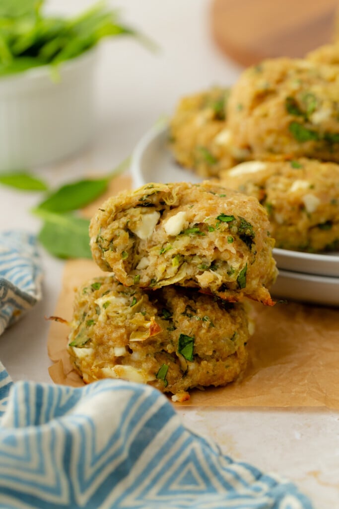 Two golden-brown veggie patties with visible spinach and cheese are stacked on parchment paper, with one broken open to show the inside. Fresh greens and a blue-striped napkin are nearby.