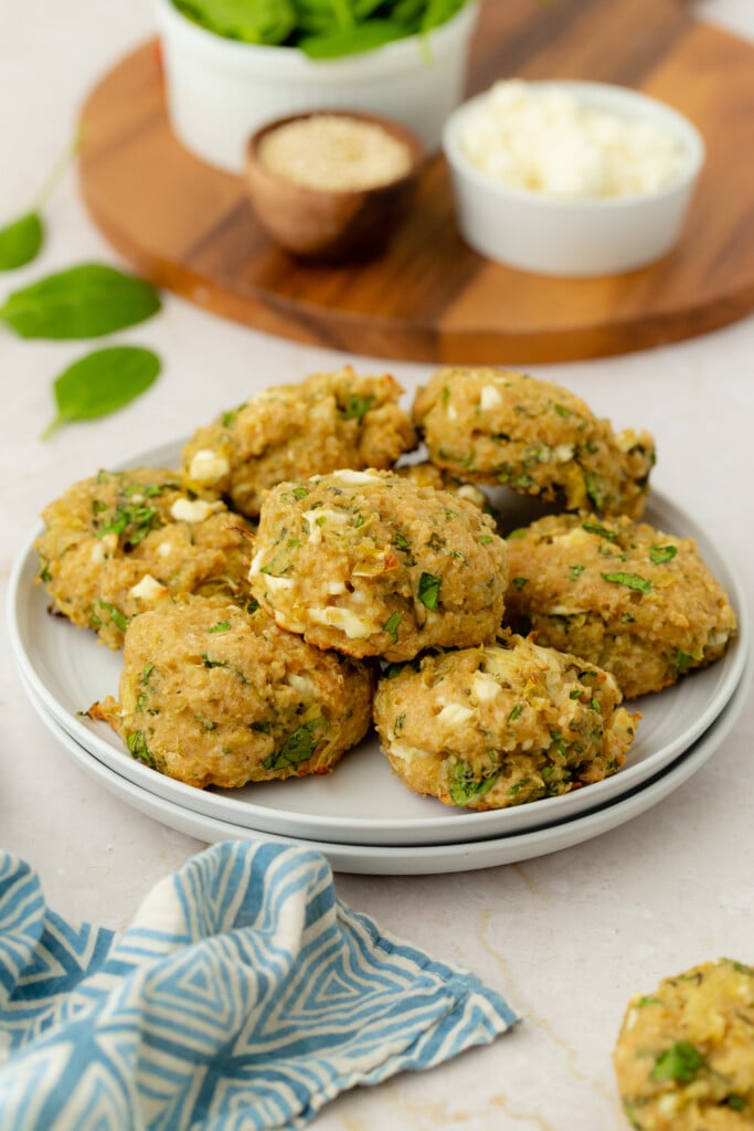 A plate of golden-brown veggie patties garnished with herbs, surrounded by a blue-striped napkin and bowls of fresh spinach, sesame seeds, and crumbled cheese on a light surface.