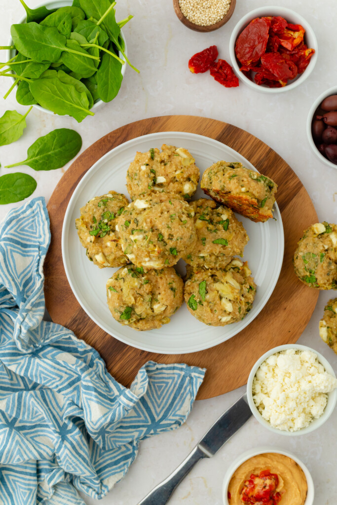 A plate of six baked falafel patties sits on a wooden board, surrounded by bowls of feta cheese, olives, sun-dried tomatoes, hummus, leafy spinach, and a blue patterned cloth napkin.