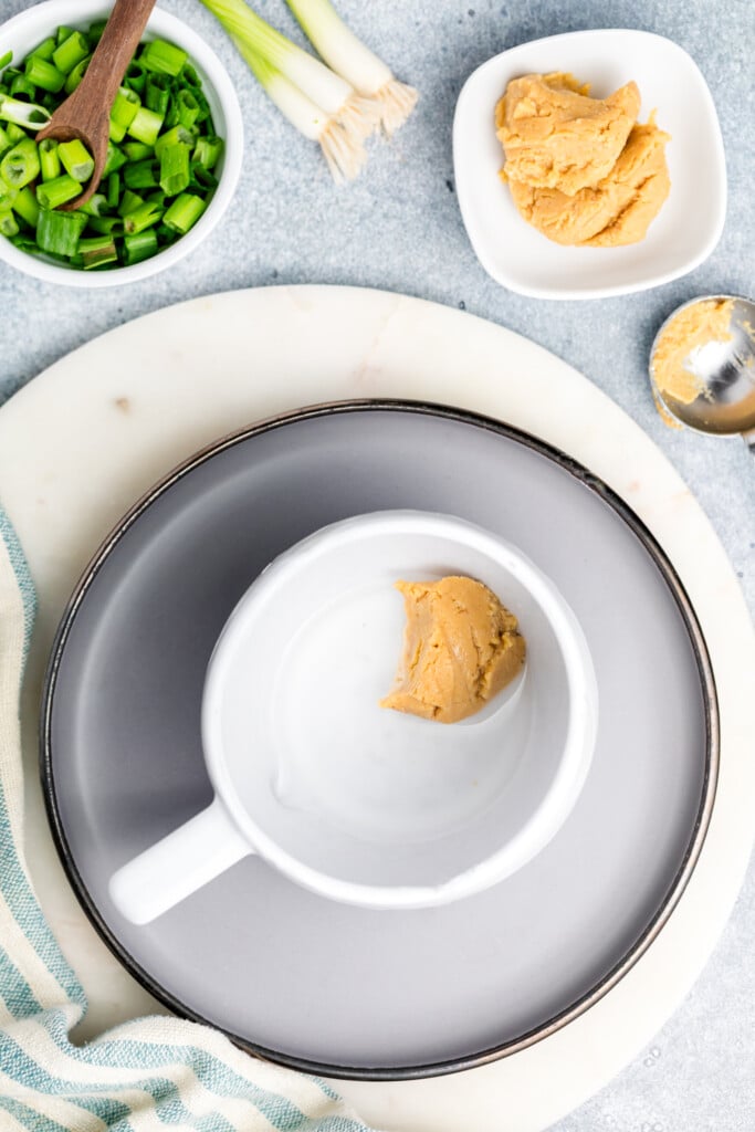 A white mug with a spoonful of miso paste inside sits on a gray plate. Nearby are bowls of chopped green onions, more miso paste, and a striped cloth on a light surface.
