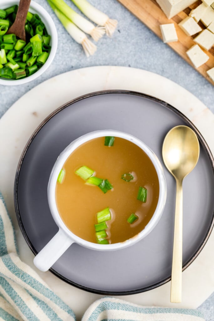 A cup of miso soup garnished with chopped green onions sits on a gray plate with a gold spoon beside it. Nearby are a bowl of green onions, a striped towel, and a cutting board with diced tofu and green onions.