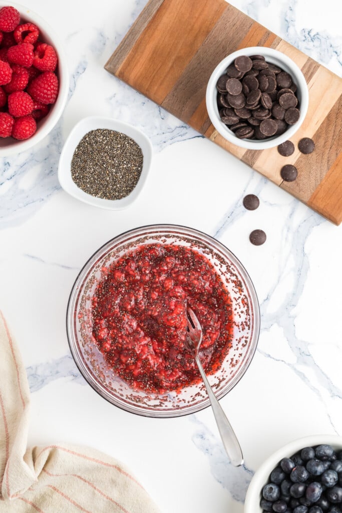 A bowl of raspberry chia jam with a spoon sits on a marble surface, surrounded by bowls of fresh raspberries, chia seeds, blueberries, and a small bowl of chocolate chips on a wooden board.