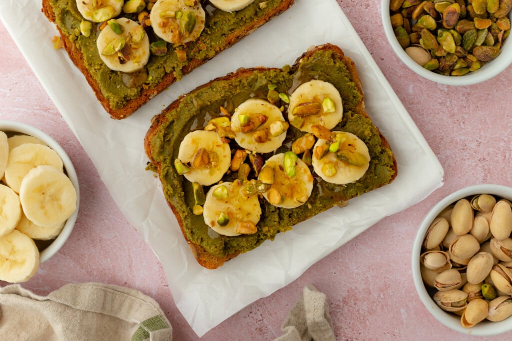 A slice of bread topped with green pistachio spread, banana slices, and chopped pistachios sits on parchment paper, surrounded by bowls of banana slices and pistachios on a pink surface.