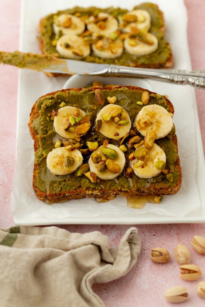A slice of toast topped with green pistachio spread, banana slices, chopped pistachios, and a drizzle of honey sits on a white plate; another similar toast and a knife are in the background.