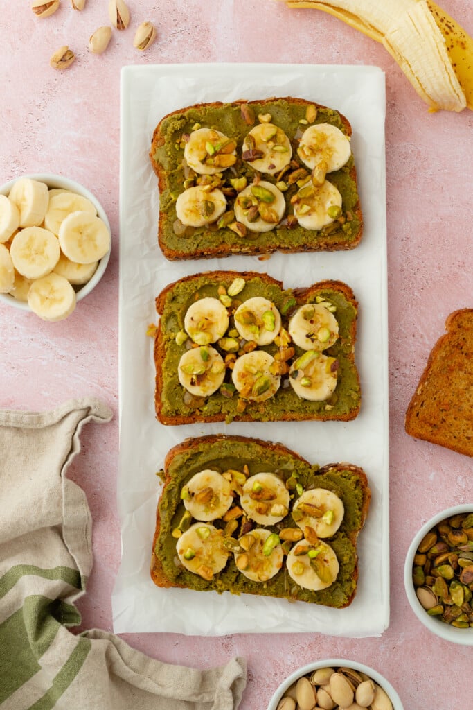 Three slices of toast topped with green spread, banana slices, chopped pistachios, and drizzle, arranged on a rectangular plate with bowls of banana slices, pistachios, and a banana nearby.