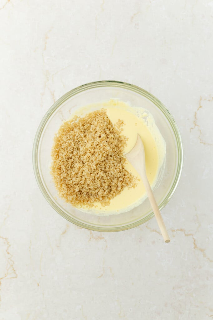 A glass bowl with a mixture of beaten eggs and light brown breadcrumbs, being stirred with a wooden spoon, sits on a light-colored marble surface.