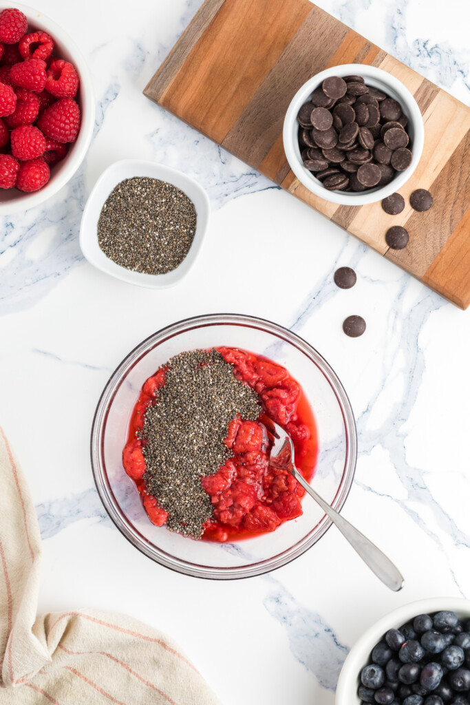 A bowl of mashed raspberries with chia seeds and a spoon on a marble countertop, surrounded by fresh raspberries, blueberries, a bowl of chocolate chips, and a bowl of chia seeds.