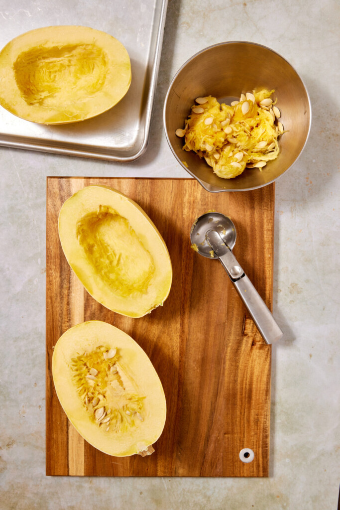 Two spaghetti squash halves on a wooden board with a scoop, another half on a baking tray, and seeds with pulp in a metal bowl are arranged on a light countertop—perfect preparation for serving Turkey Meatballs with Spaghetti Squash.
