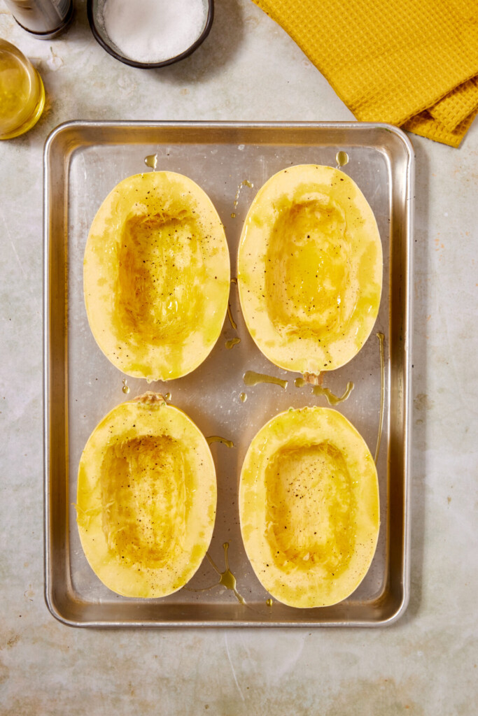 Four halved spaghetti squash with seeds removed are arranged cut side up on a baking sheet. They are drizzled with oil, sprinkled with pepper, and ready for roasting—the perfect base for Turkey Meatballs with Spaghetti Squash. A yellow towel and small bowl are nearby.