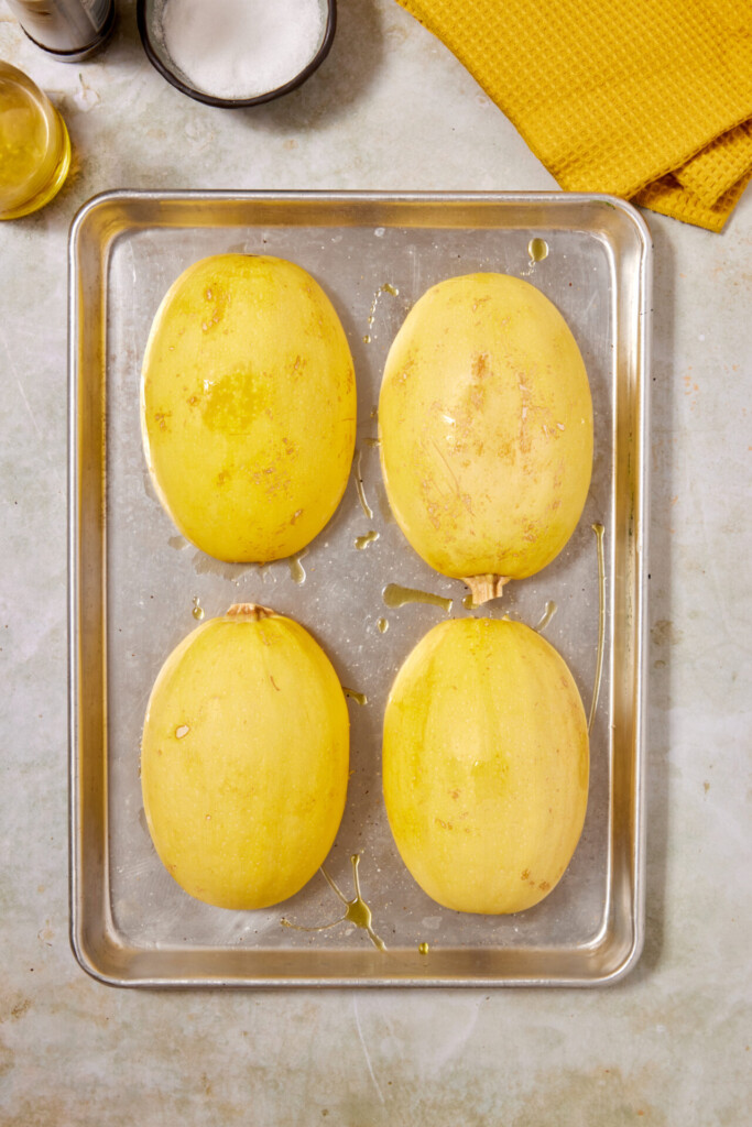 Four halved spaghetti squashes, cut side down, are placed on a baking sheet with a drizzle of oil—perfectly prepped for Turkey Meatballs with Spaghetti Squash. A yellow cloth, a bowl of salt, and a bottle of olive oil are nearby.