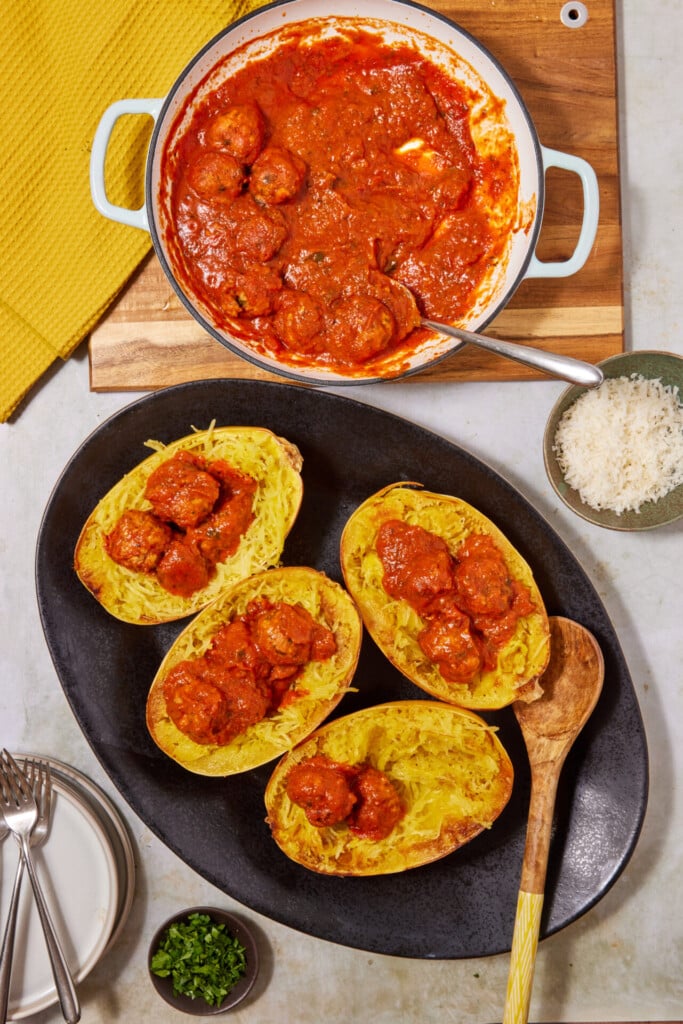 Four spaghetti squash halves filled with savory tomato sauce and turkey meatballs on a black platter, next to a pot of extra sauce and bowls of grated cheese and chopped herbs, with plates and forks nearby—perfect for serving up Turkey Meatballs with Spaghetti Squash.