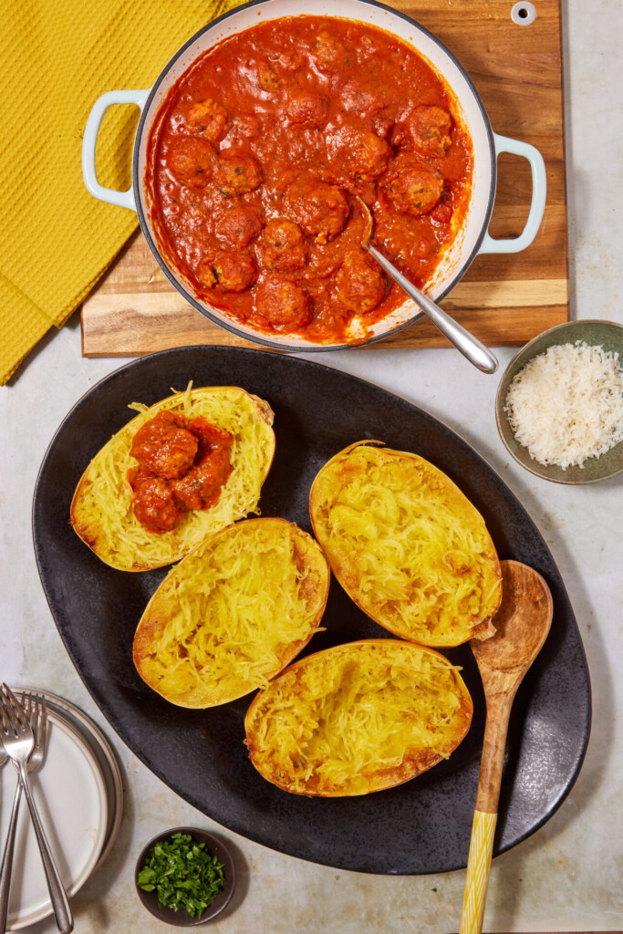 Baked spaghetti squash halves on a black platter, one topped with savory turkey meatballs in tomato sauce. A pot of Turkey Meatballs with Spaghetti Squash, grated cheese, chopped herbs, plates, forks, and a yellow napkin are nearby.