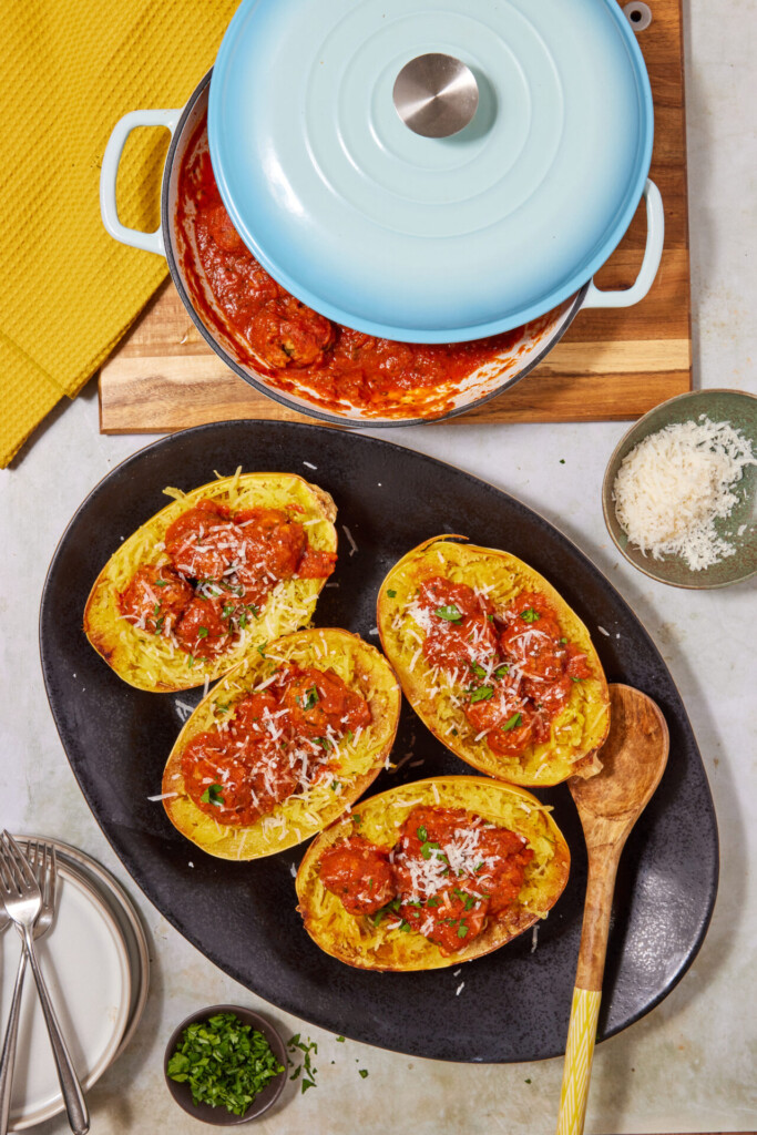 Four spaghetti squash halves topped with tomato sauce and grated cheese on a black tray, accompanied by savory Turkey Meatballs with Spaghetti Squash, with a blue pot of sauce, grated cheese bowl, chopped herbs, plates, forks, and a yellow towel nearby.