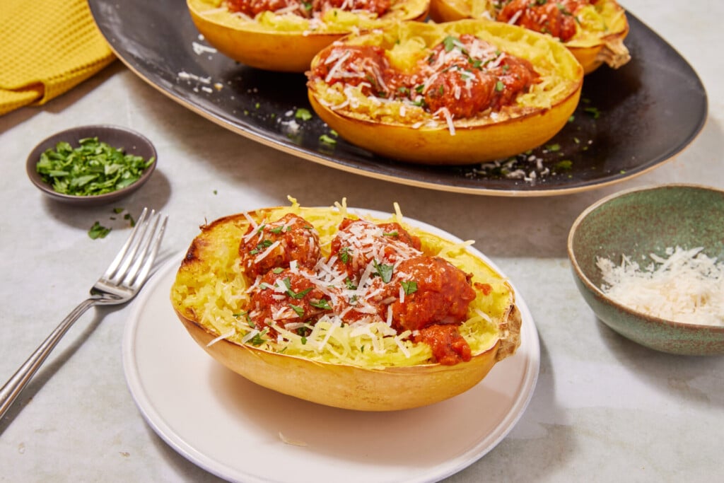 A spaghetti squash half filled with tender spaghetti squash strands and savory turkey meatballs in tomato sauce, topped with grated cheese, served on a white plate with a fork, alongside extra cheese and fresh herbs for garnish. Enjoy this wholesome Turkey Meatballs with Spaghetti Squash dish for a satisfying and delicious meal.