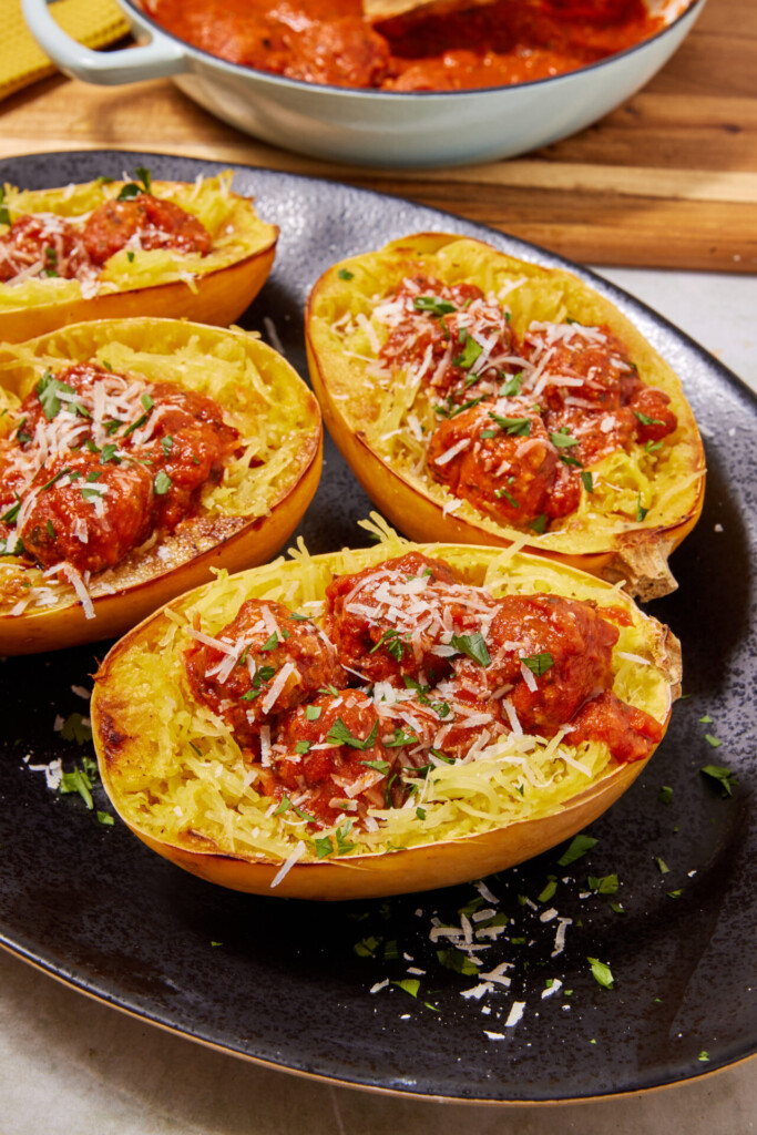 Four spaghetti squash halves on a black plate, filled with turkey meatballs in tomato sauce and sprinkled with grated cheese and herbs. A pan of extra Turkey Meatballs with Spaghetti Squash sits in the background.