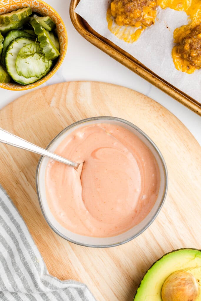 A bowl of creamy pink sauce with a spoon, next to sliced pickles, baked cheesy meatballs on a tray, a halved avocado, and a striped cloth on a wooden board.