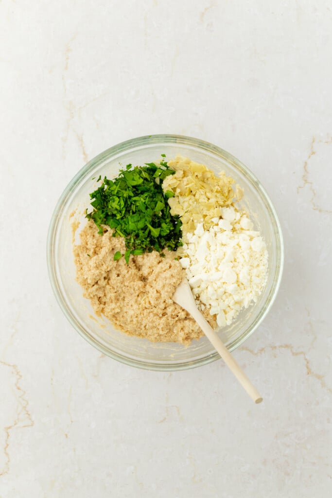 A glass bowl on a light marble surface contains chopped parsley, crumbled feta cheese, chopped artichoke hearts, and a beige hummus-like mixture, with a wooden spoon resting inside.