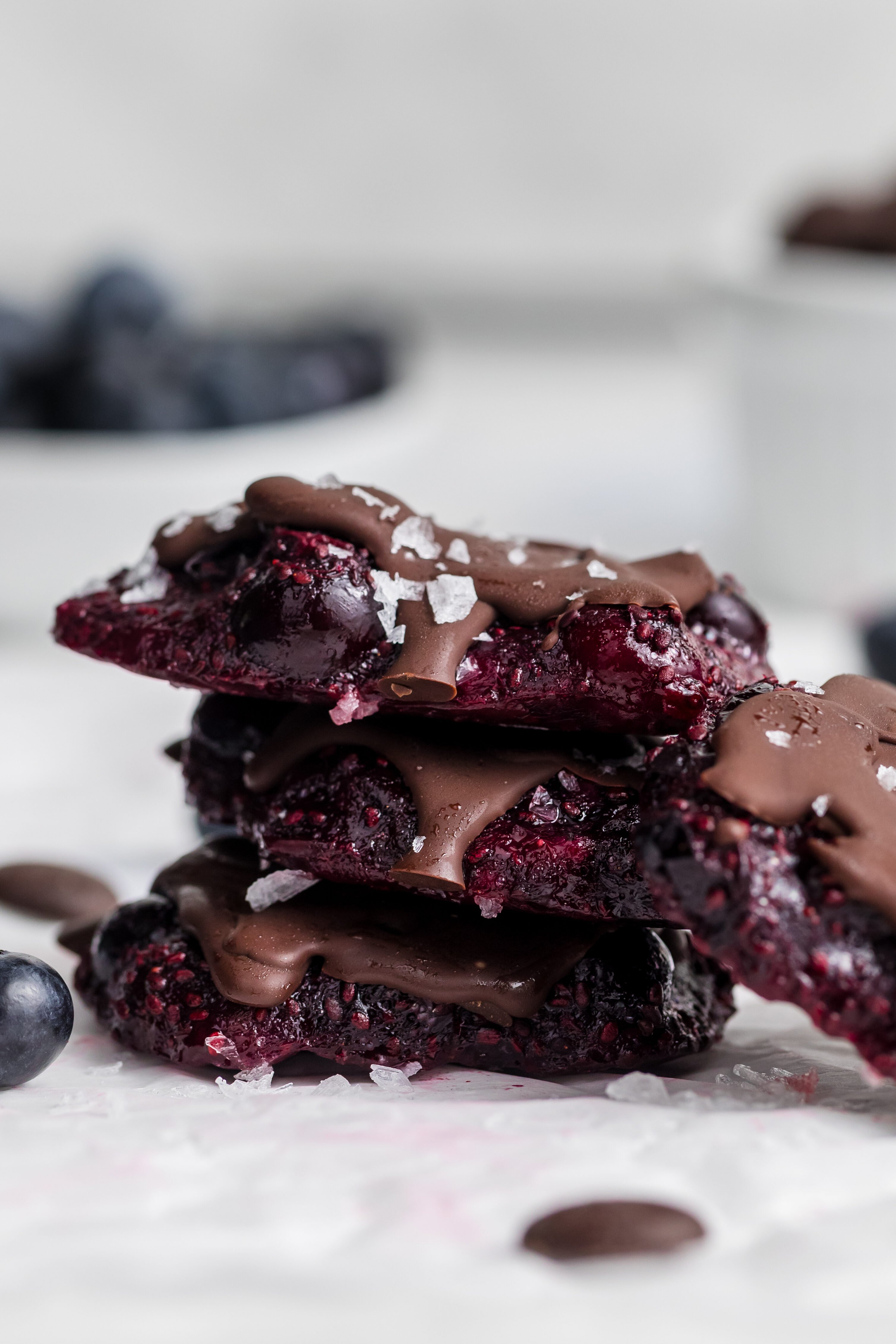 Three stacked pieces of dark chocolate and berry bark drizzled with chocolate and topped with flaky salt, surrounded by fresh blueberries and chocolate chips, with bowls of blueberries and chocolate in the background.