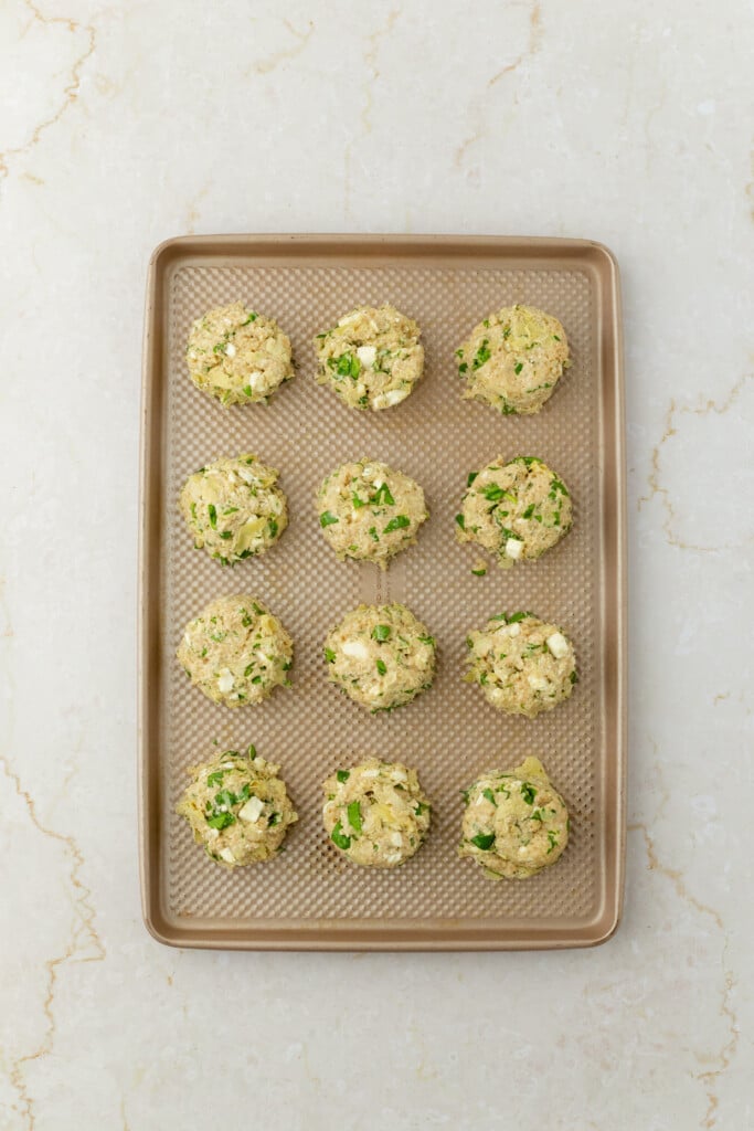 A baking sheet with twelve unbaked, round falafel balls containing visible green herbs, placed evenly on a light-colored textured surface.