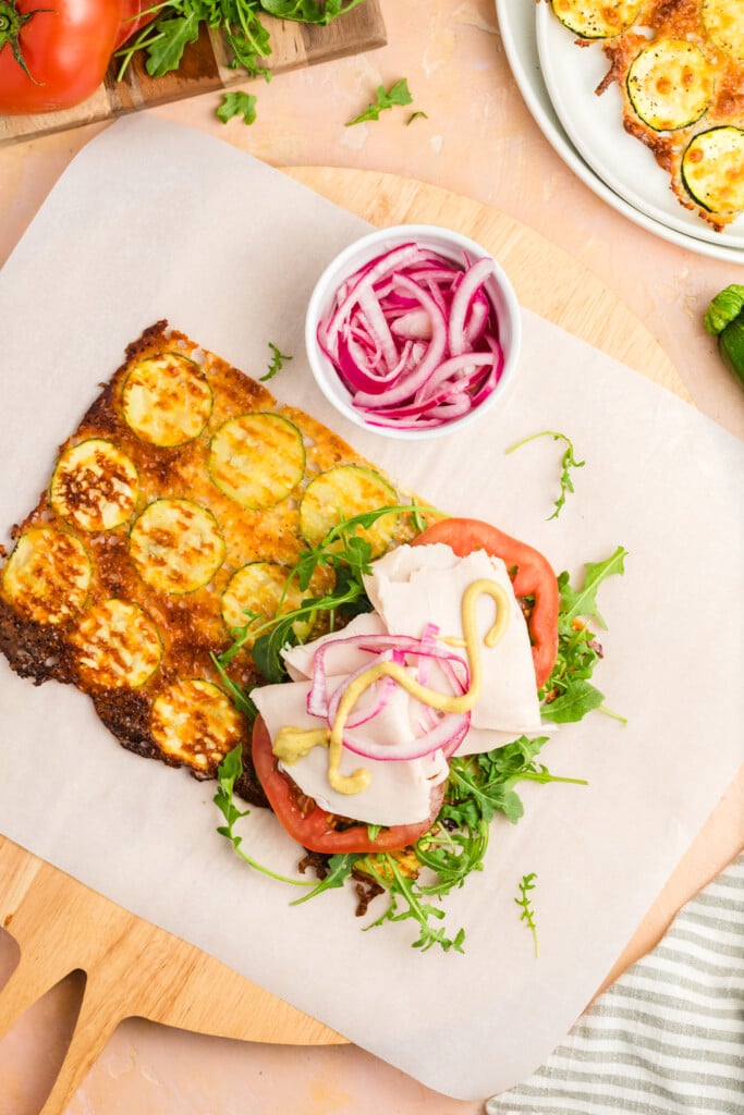 A slice of Zucchini Cheese Flatbread topped with arugula, tomato, turkey, avocado, mustard, and pickled onions sits on parchment paper beside a bowl of pickled onions. Sliced zucchini and fresh greens complete the vibrant scene.