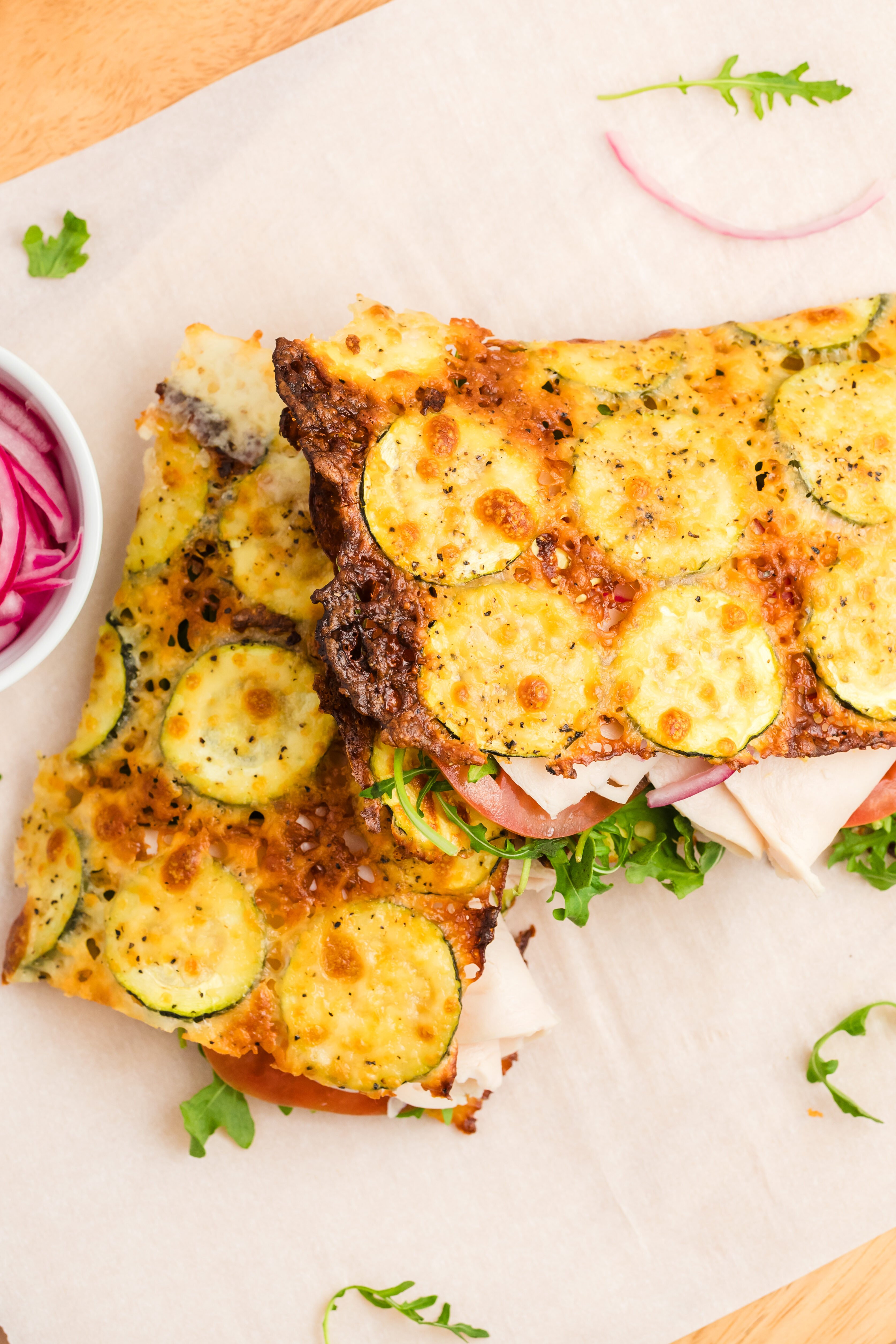 Two open-faced zucchini cheese flatbread sandwiches on parchment paper, topped with melted cheese, zucchini slices, turkey, tomato, and arugula, with a small bowl of sliced red onions on the side.