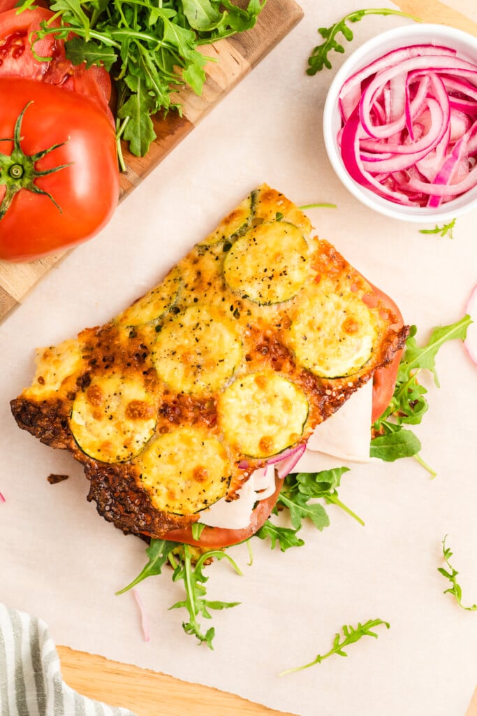 Open-faced sandwich with arugula, tomato, red onion, and deli meat, topped with a slice of Zucchini Cheese Flatbread. Fresh tomato, arugula, and a bowl of sliced red onions rest on a parchment-covered surface.