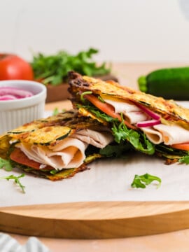 Two stacked sandwiches with turkey, arugula, tomato, and red onion between thin, grilled Zucchini Cheese Flatbread, displayed on a wooden board with fresh vegetables and pickled onions nearby.