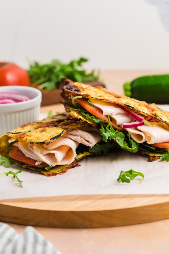 Two stacked sandwiches with turkey, arugula, tomato, and red onion between thin, grilled Zucchini Cheese Flatbread, displayed on a wooden board with fresh vegetables and pickled onions nearby.