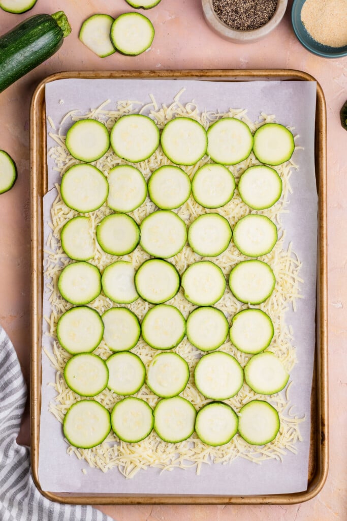 A baking sheet lined with parchment paper holds a single layer of zucchini slices arranged on top of shredded cheese, ready to become Zucchini Cheese Flatbread. A striped towel, fresh zucchini, and bowls of ingredients are nearby.