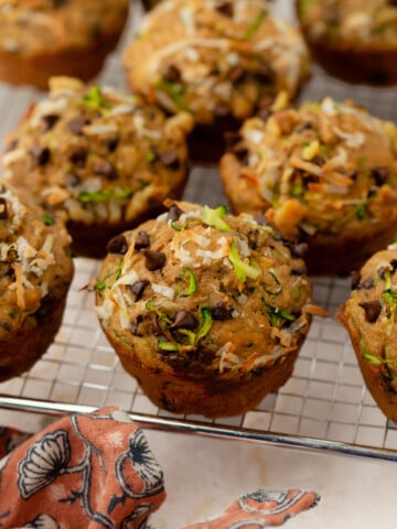 A close-up of freshly baked zucchini muffins with visible shredded zucchini, chocolate chips, and coconut flakes, cooling on a wire rack next to a patterned cloth.