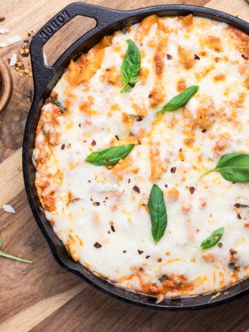 A cast iron skillet filled with baked cheesy pasta topped with melted cheese, fresh basil leaves, and sprinkled red pepper flakes. The skillet is on a wooden board with a small bowl of red pepper flakes and basil nearby.