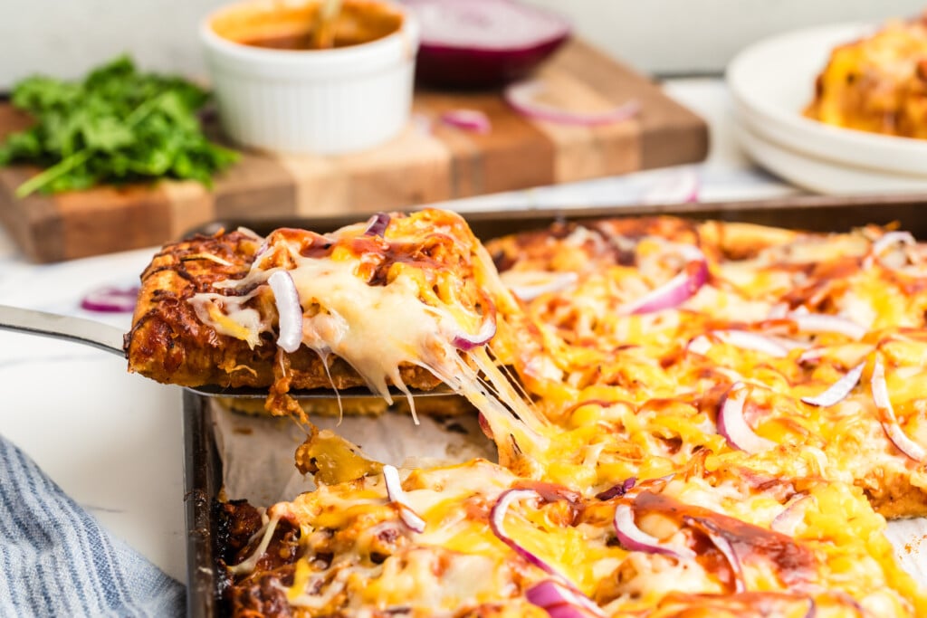 A metal spatula lifts a cheesy, golden-brown slice of pizza from a baking sheet. The pizza has melted cheese, red onion slices, and barbecue sauce. Fresh parsley and sauce are blurred in the background.